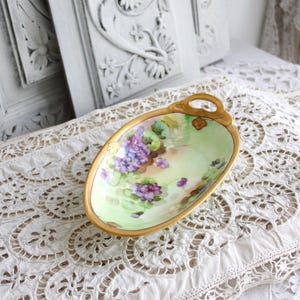 May include: A vintage oval-shaped serving dish with a green and white floral design featuring purple violets. The dish has two gold handles and a gold rim.