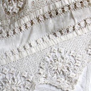 May include: Detailed close-up of a white lace textile. The lace displays a variety of patterns, including floral and geometric designs. The texture is intricate, with visible stitching and openwork, giving a delicate and elegant look. The textile appears to be a tablecloth or similar decorative item.