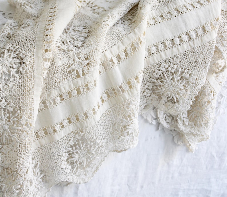 May include: Close-up of a cream-coloured lace textile. The fabric displays detailed patterns, including floral and geometric designs. The lace is draped, highlighting its texture and delicate craftsmanship. The overall aesthetic is vintage and elegant.