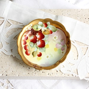 May include: A vintage porcelain bowl with a gold rim and a hand-painted design of strawberries and flowers. The bowl is ideal for serving fruit or sweets.