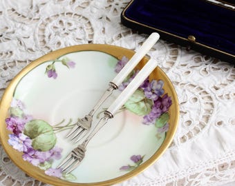 Może przedstawiać: Zabytkowy porcelanowy talerz ze złotą obwódką i ręcznie malowanym fioletowym wzorem kwiatowym, w towarzystwie dwóch małych widelców z białymi uchwytami. Talerz spoczywa na białym koronkowej obrusie, z ciemnoniebieskim, aksamitnym etui w tle.