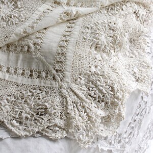 May include: A close-up of a cream-coloured, vintage lace textile. The intricate design features floral patterns and openwork details. The lace is draped over a white surface, showcasing its delicate texture and craftsmanship. The textile appears to be in good condition.