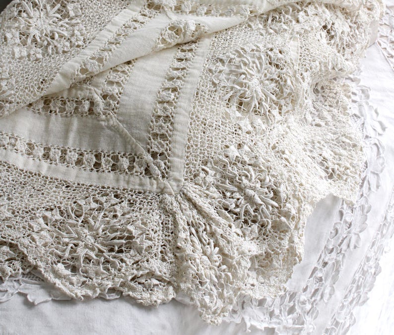 May include: A close-up of a cream-coloured, vintage lace textile. The intricate design features floral patterns and openwork details. The lace is draped over a white surface, showcasing its delicate texture and craftsmanship. The textile appears to be in good condition.