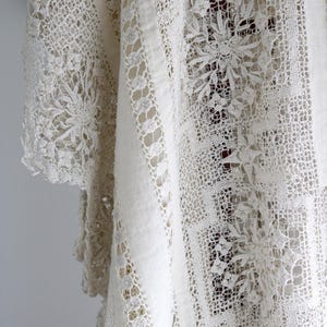 May include: A close-up view of a cream-coloured vintage lace textile. The fabric displays detailed floral and geometric patterns, with solid and openwork sections. The lace is draped, highlighting the texture and design details.