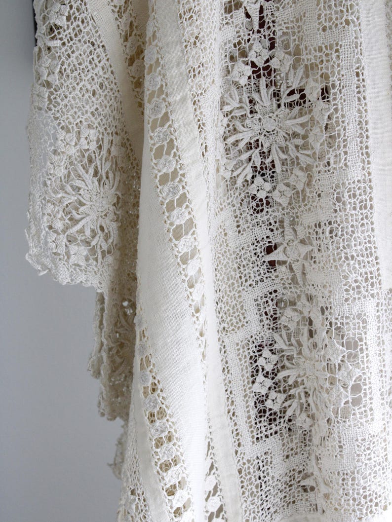 May include: A close-up view of a cream-coloured vintage lace textile. The fabric displays detailed floral and geometric patterns, with solid and openwork sections. The lace is draped, highlighting the texture and design details.