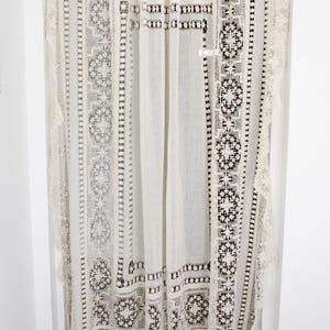 May include: Cream-coloured lace curtain with intricate floral and geometric patterns. The curtain has a scalloped edge and is hung from a rod. The design includes detailed openwork and borders, creating a delicate and elegant aesthetic.