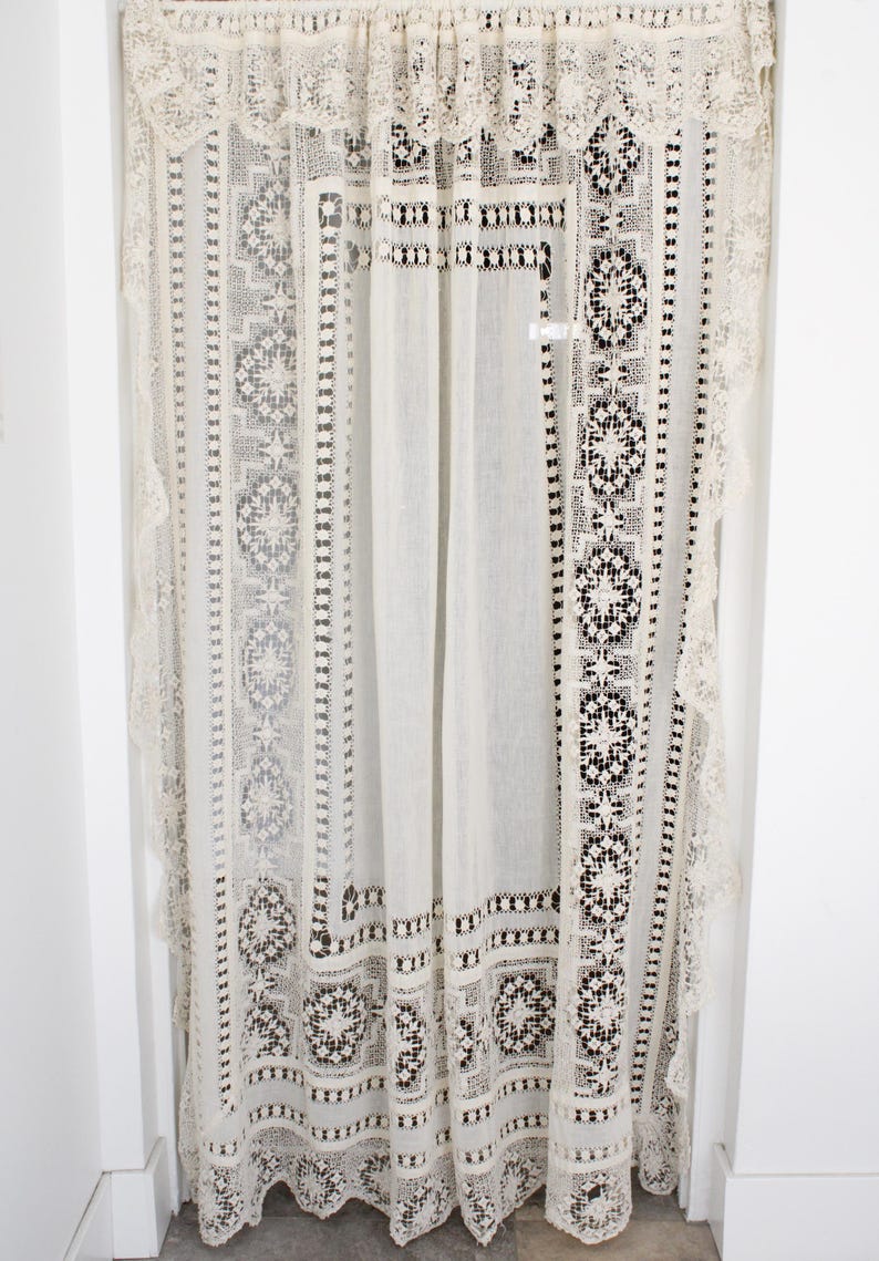 May include: Cream-coloured lace curtain with intricate floral and geometric patterns. The curtain has a scalloped edge and is hung from a rod. The design includes detailed openwork and borders, creating a delicate and elegant aesthetic.
