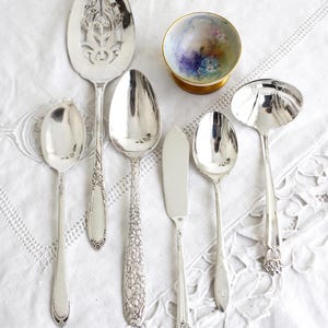 Könnte beinhalten: Eine Sammlung antiker silberner Servierutensilien, darunter Löffel, ein Buttermesser und eine Schöpfkelle, auf einer weißen Spitzendecke angeordnet. Eine kleine, dekorative Schale mit floralem Design ist ebenfalls vorhanden.