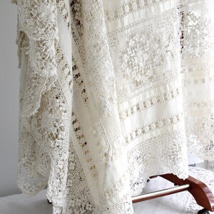 May include: Cream-coloured lace tablecloth with intricate openwork patterns. The vintage textile features detailed floral and geometric designs, draped over a wooden stand. The lace has a delicate, textured appearance, showcasing the craftsmanship.