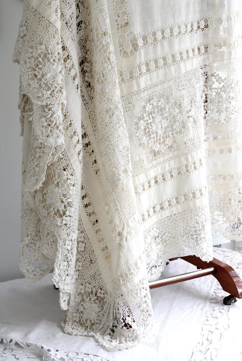 May include: Cream-coloured lace tablecloth with intricate openwork patterns. The vintage textile features detailed floral and geometric designs, draped over a wooden stand. The lace has a delicate, textured appearance, showcasing the craftsmanship.
