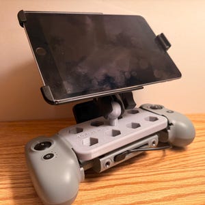 May include: A gray tablet holder with a black tablet attached. The holder is mounted on a gray game controller with two gray handles. The tablet is held in place by black clips. The holder is on a wooden surface.