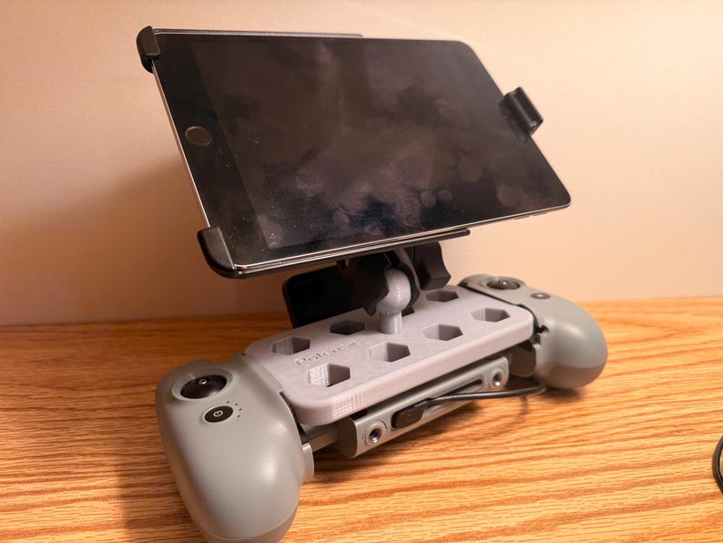 May include: A gray tablet holder with a black tablet attached. The holder is mounted on a gray game controller with two gray handles. The tablet is held in place by black clips. The holder is on a wooden surface.