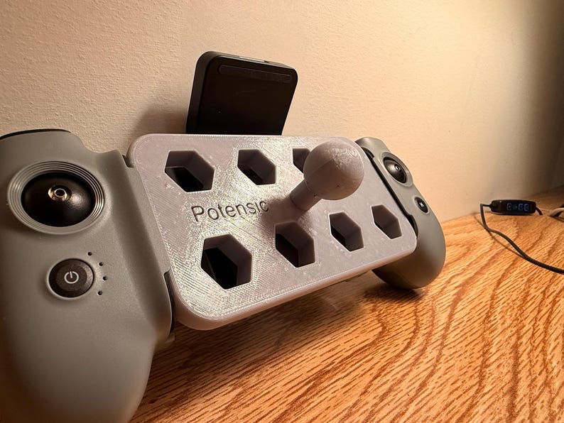 May include: A gray drone controller with a 3D-printed gray holder attached. The holder has a joystick and the word "Potensic" printed on it. A black device is mounted on the holder.