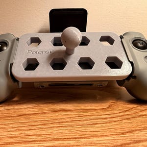 May include: A gray drone controller with the brand name "Potensic" printed on the front. The controller has a central platform with hexagonal cutouts and a joystick. It features two side grips with buttons and a black phone holder.