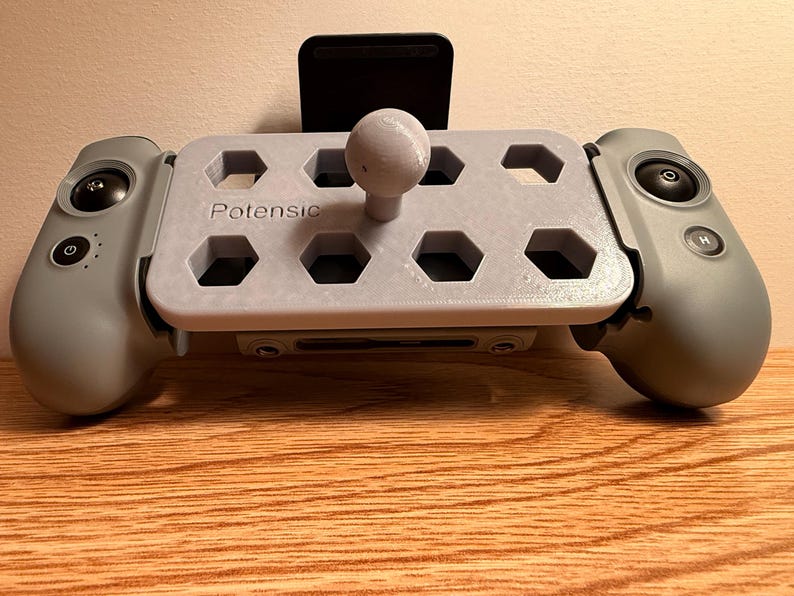 May include: A gray drone controller with the brand name "Potensic" printed on the front. The controller has a central platform with hexagonal cutouts and a joystick. It features two side grips with buttons and a black phone holder.