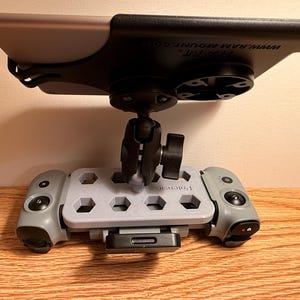 May include: A gray and black tablet mount with a ball joint and a base with hexagonal cutouts. The mount has a black top and a gray base with the word "Patensic" printed on it. The mount is designed to hold a tablet.