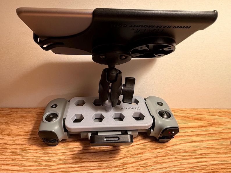May include: A gray and black tablet mount with a ball joint and a base with hexagonal cutouts. The mount has a black top and a gray base with the word "Patensic" printed on it. The mount is designed to hold a tablet.