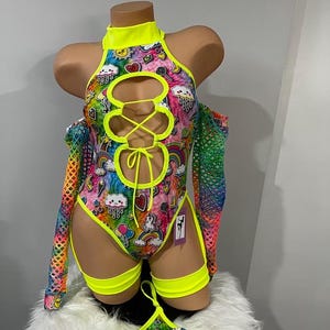Puede incluir: Un body de color amarillo neón y tie-dye arcoíris con un frente con cordones y un estampado gráfico de unicornio, arcoíris y corazón. El body incluye ligas amarillas neón a juego y un tanga de color amarillo neón y tie-dye arcoíris a juego.