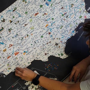 Person pinning a detailed USA travel map. Highlighting  landmarks and national parks, this USA Map has images ot top attractions visible for each state. The USA Map is printed on canvas and framed. The background is charcoal, the land is light beige.