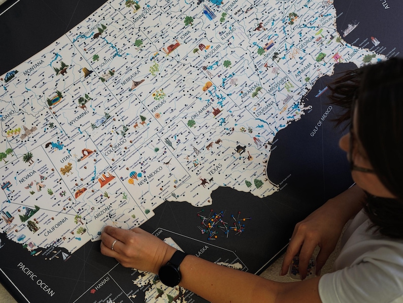 Person pinning a detailed USA travel map. Highlighting  landmarks and national parks, this USA Map has images ot top attractions visible for each state. The USA Map is printed on canvas and framed. The background is charcoal, the land is light beige.