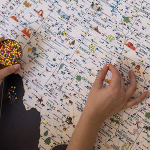 Hands placing push pins on a USA travel map. Customizable and framed, this USA map highlights state landmarks, national parks, and travel routes. Drawings with top touristic attractions are visible.
