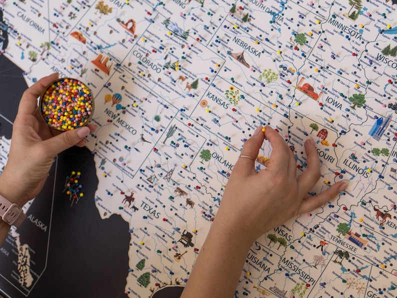 Hands placing push pins on a USA travel map. Customizable and framed, this USA map highlights state landmarks, national parks, and travel routes. Drawings with top touristic attractions are visible.