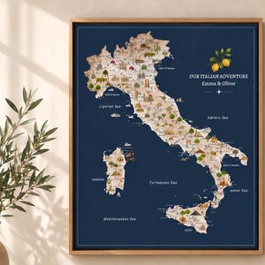 May include: A framed wall art print featuring a map of Italy on a dark blue background. The map displays various landmarks and illustrations across the country. Text reads: OUR ITALIAN ADVENTURE, Emma & Oliver. Two yellow lemons are depicted above the text.