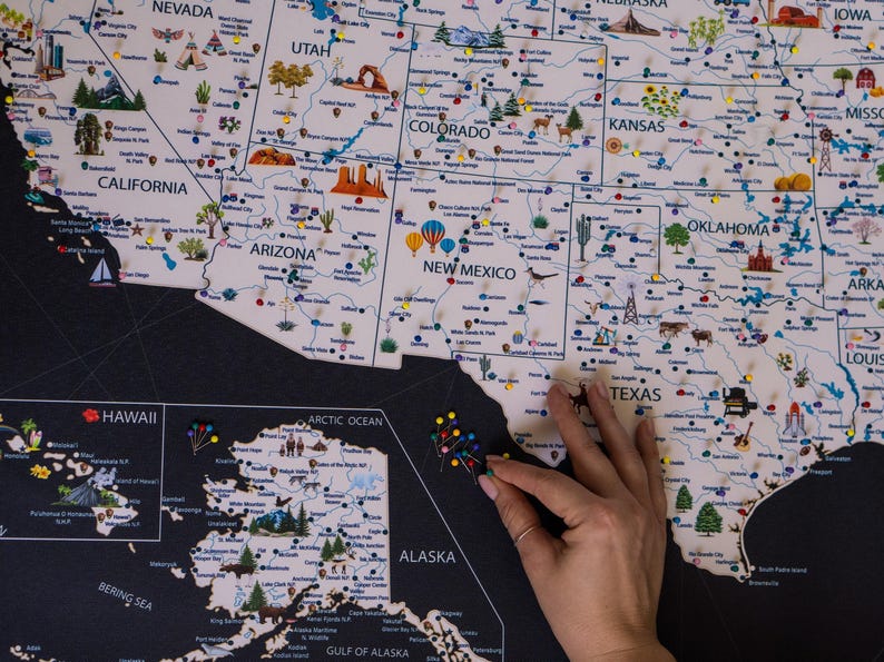 A close up with the USA Map with top attractions, landmarks, national parks and cities. In the center, a hand is holding a push pin, to place it on the USA Map. Printed on canvas, the background of the USA Map is charcoal and the land is light beige.
