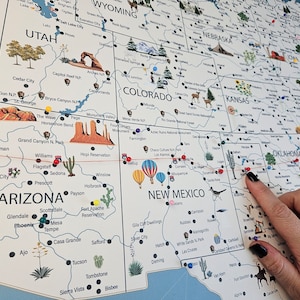May include: A close-up of a map of the United States with red push pins marking locations. The map is on a white background with black lines and text. A hand with black nail polish is pointing to a red push pin in New Mexico.