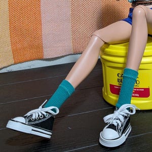 Könnte beinhalten: Eine Puppe mit blauen Shorts, gerippten türkisfarbenen Socken und schwarz-weißen Schnürschuhen. Die Puppe sitzt auf einem gelben Kunststoffbehälter mit der Aufschrift Cafe Bustelo, Nettogewicht 283g, mit einem roten Streifen und weißem Text.