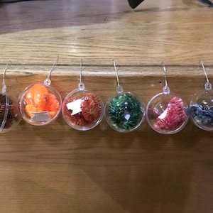 May include: Six clear Christmas ornaments with various colorful fillings, including an orange octopus, and yarn in shades of brown, red, green, and blue. Each ornament has a silver hook for hanging. The ornaments are displayed on a wooden surface.