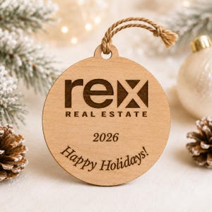 Könnte beinhalten: Ein runder, hellfarbiger Holzanhänger mit der lasergravierten Aufschrift "rex REAL ESTATE", dem Jahr "2026" und dem Schriftzug "Happy Holidays!" in dunkelbrauner Farbe. Der Anhänger besitzt eine Schlaufe aus natürlicher Jutekordel.