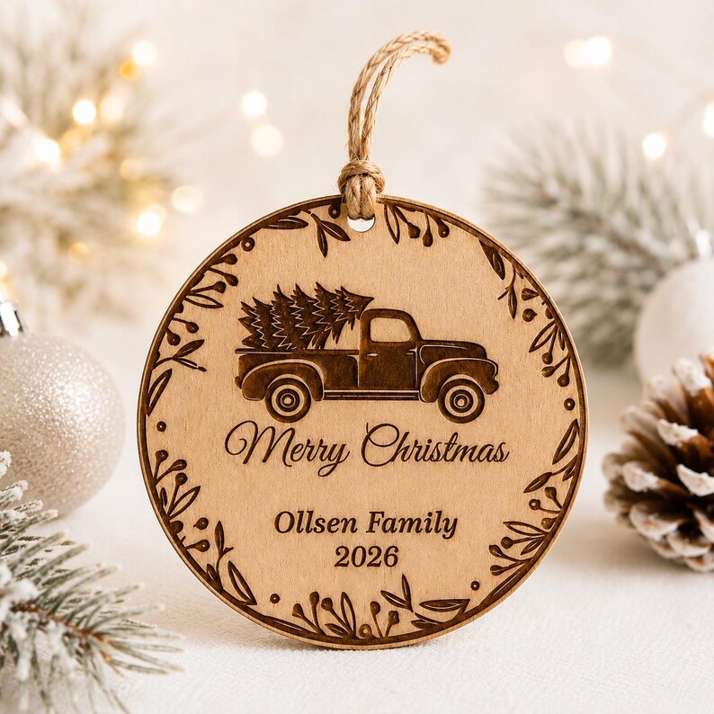 K&ouml;nnte beinhalten: Ein runder Holzanh&auml;nger mit einer Lasergravur eines alten Pick-ups, der einen Weihnachtsbaum transportiert. Der Text lautet Merry Christmas, Ollsen Family und 2026. Ein dekorativer Blattrand umrahmt das Motiv, erg&auml;nzt durch eine Juteschnur.