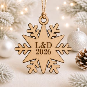 K&ouml;nnte beinhalten: Ein helles Holzornament in Form einer Schneeflocke mit einer Lasergravur der Buchstaben L&D und der Jahreszahl 2026 in der Mitte. Das Ornament ist an einer d&uuml;nnen, hellbraunen Schnur befestigt, die durch eine runde &Ouml;ffnung oben gef&uuml;hrt wird.