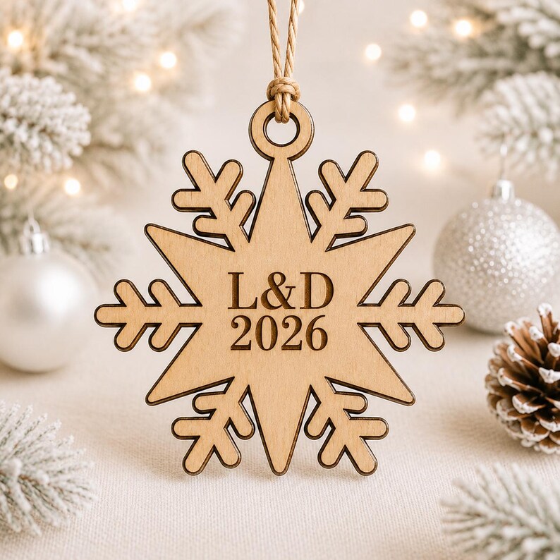 K&ouml;nnte beinhalten: Ein helles Holzornament in Form einer Schneeflocke mit einer Lasergravur der Buchstaben L&D und der Jahreszahl 2026 in der Mitte. Das Ornament ist an einer d&uuml;nnen, hellbraunen Schnur befestigt, die durch eine runde &Ouml;ffnung oben gef&uuml;hrt wird.