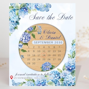 May include: A wedding invitation with a watercolour design. The invitation features a wooden calendar with the date circled, surrounded by blue hydrangea flowers. The text "Save the Date" and names are displayed at the top.