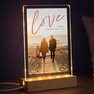 以下が含まれることがあります： 筆記体で「love」と書かれた、光るフォトフレーム。夕暮れのビーチの木製遊歩道を歩くカップルの画像。テキスト「Happy Valentine's Day」も表示されています。フレームは木製の台座に置かれています。