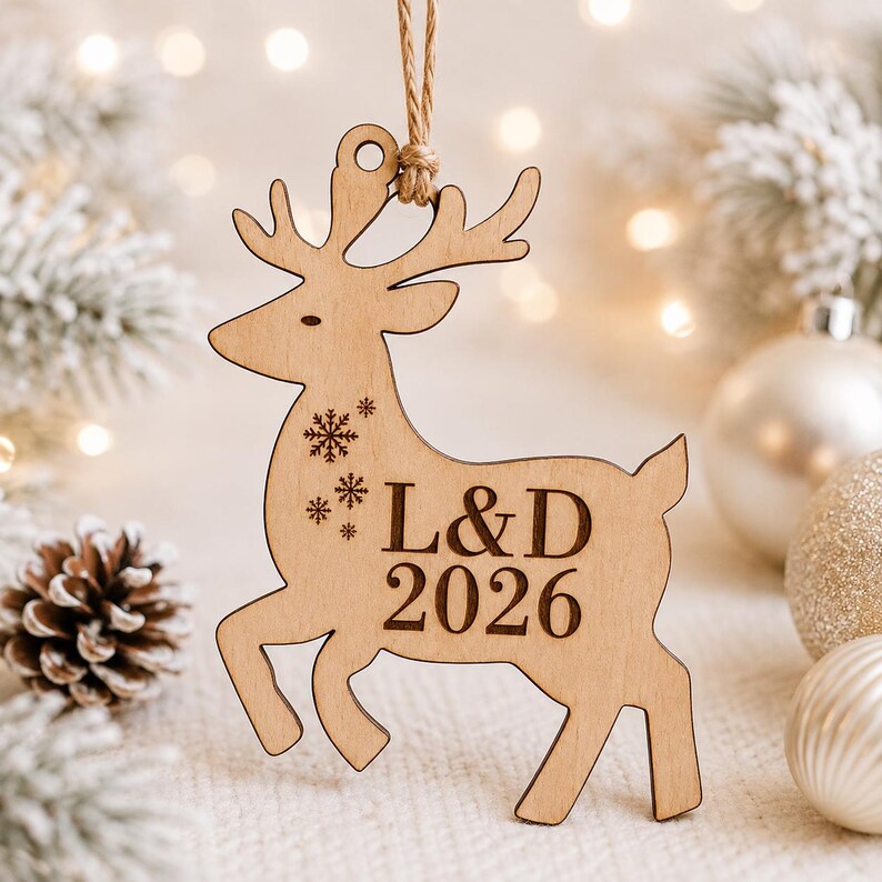 K&ouml;nnte beinhalten: Ein helles Holzornament in Rentierform mit Ausschnittdesign, das an einer Juteschnur h&auml;ngt. Das Rentier ist mit drei kleinen Schneeflockensymbolen und dem Text L&D 2026 in einer dunklen Serifenschrift auf der Seite graviert.