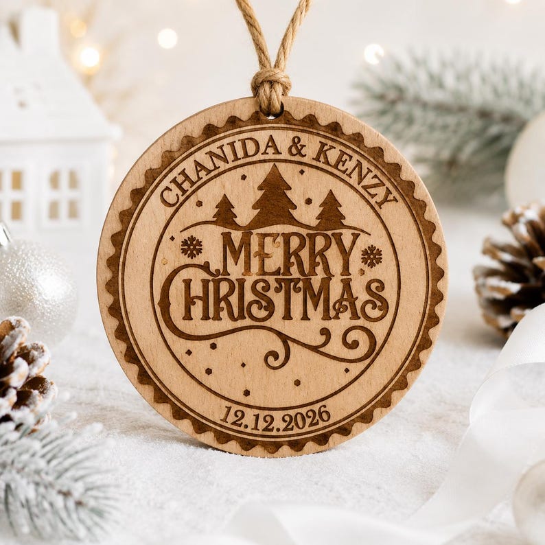 K&ouml;nnte beinhalten: Ein runder Weihnachtsanh&auml;nger aus Holz mit gewelltem Rand, laser-graviert mit dem Text CHANIDA & KENZY, MERRY CHRISTMAS und dem Datum 12.12.2026, verziert mit Tannenbaum- und Schneeflockenmotiven, aufgeh&auml;ngt an einer beigen Kordelschlaufe.