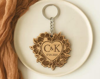 Könnte beinhalten: Ein Holzschlüsselanhänger mit floralem Design und herzförmiger Mitte. Das Herz enthält die Initialen "C&K" und das Datum "17-05-2025". Der Schlüsselanhänger ist an einem silbernen Metallring befestigt.