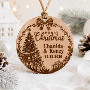 K&ouml;nnte beinhalten: Ein runder Weihnachtsanh&auml;nger aus Holz mit einer Lasergravur eines geschm&uuml;ckten Baumes, Geschenken und einer Schleife. Der Text lautet Merry Christmas, Chanida & Kenzy, und 12.12.2026. Der Anh&auml;nger h&auml;ngt an einer rustikalen Kordelschlaufe.