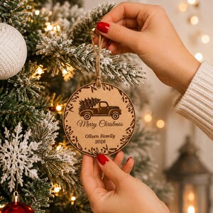 K&ouml;nnte beinhalten: Ein runder Weihnachtsbaumschmuck aus hellem Holz mit einer Lasergravur eines Pick-ups, der einen Tannenbaum transportiert. Der Text Merry Christmas, Ollsen Family und 2026 ist unter dem Fahrzeug eingraviert, umrahmt von einem Blattmuster.