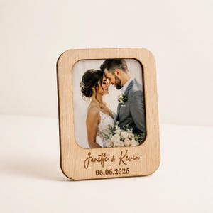Puede incluir: Un marco de fotos de madera con esquinas redondeadas, que muestra una foto de boda de una pareja. El marco está grabado con los nombres "Janette & Kevin" y la fecha "06.06.2026". La foto muestra a la pareja abrazándose, con la novia vestida de blanco.