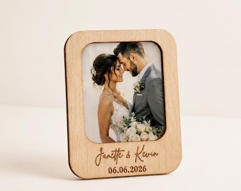 Könnte beinhalten: Ein rechteckiger Bilderrahmen aus hellem Holz mit einem Foto eines sich umarmenden Paares. Der Rahmen ist im unteren Bereich mit den Namen Janette & Kevin sowie dem Datum 06.06.2026 in einer kursiven Schriftart graviert.