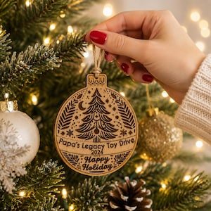 K&ouml;nnte beinhalten: Ein runder Weihnachtsbaumschmuck aus Holz mit einer lasergravierten Tanne, einer Mondsichel und Blattmotiven. Der Text lautet Papa's Legacy Toy Drive 2024 Happy Holiday. Der Anh&auml;nger ist an einer beigen Kordel befestigt und h&auml;ngt am Baum.