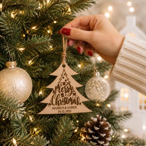 K&ouml;nnte beinhalten: Ein hellfarbiger Weihnachtsbaumschmuck aus Holz mit einer lasergravierten Darstellung eines kleinen Baumes, dem Schriftzug "Merry Christmas" sowie den Namen "MARKUS & LOREN" und dem Datum "30.12.2022", h&auml;ngend an einer braunen Kordel.