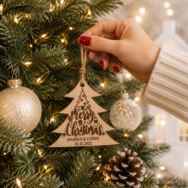 K&ouml;nnte beinhalten: Ein hellfarbiger Weihnachtsbaumschmuck aus Holz mit einer lasergravierten Darstellung eines kleinen Baumes, dem Schriftzug "Merry Christmas" sowie den Namen "MARKUS & LOREN" und dem Datum "30.12.2022", h&auml;ngend an einer braunen Kordel.