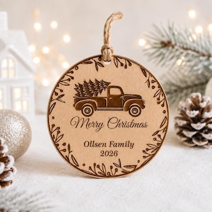 K&ouml;nnte beinhalten: Ein runder Holzanh&auml;nger mit Juteschnur, der einen lasergravierten Oldtimer-Pickup mit Weihnachtsbaum zeigt. Der Text lautet Merry Christmas, Ollsen Family und 2026, umrahmt von einer dekorativen Bord&uuml;re aus Bl&auml;ttern.
