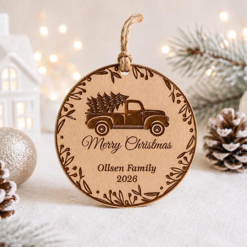 K&ouml;nnte beinhalten: Ein runder Holzanh&auml;nger mit Juteschnur, der einen lasergravierten Oldtimer-Pickup mit Weihnachtsbaum zeigt. Der Text lautet Merry Christmas, Ollsen Family und 2026, umrahmt von einer dekorativen Bord&uuml;re aus Bl&auml;ttern.