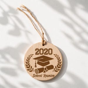 May include: A circular wooden ornament featuring a graduation cap, scroll, and the year 2020, suspended by twine. The words "School Reunion" are engraved below. The ornament is a light brown color.